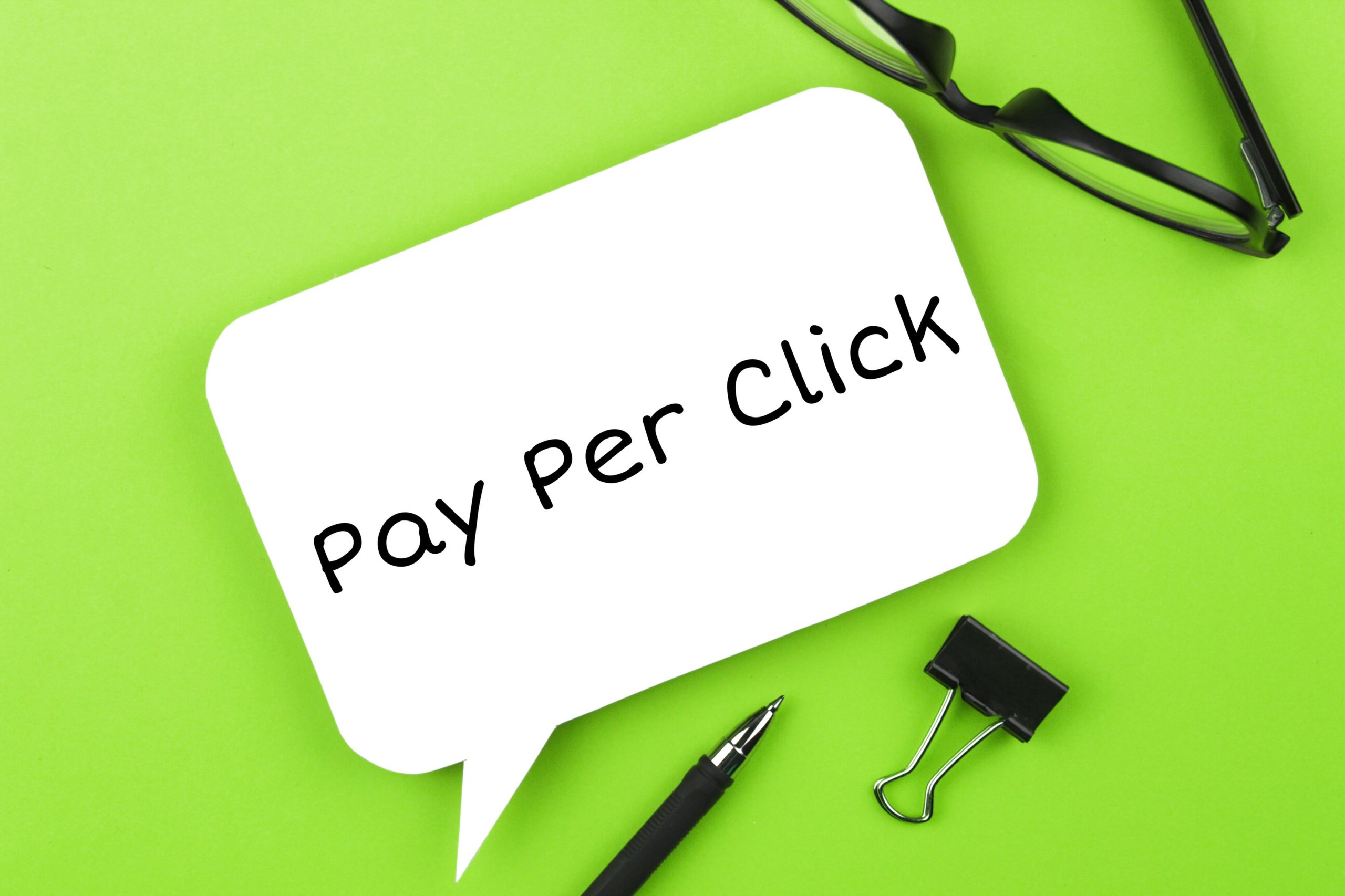 Pay Per Click concept with speech bubble, glasses, pen, and clip on green background.