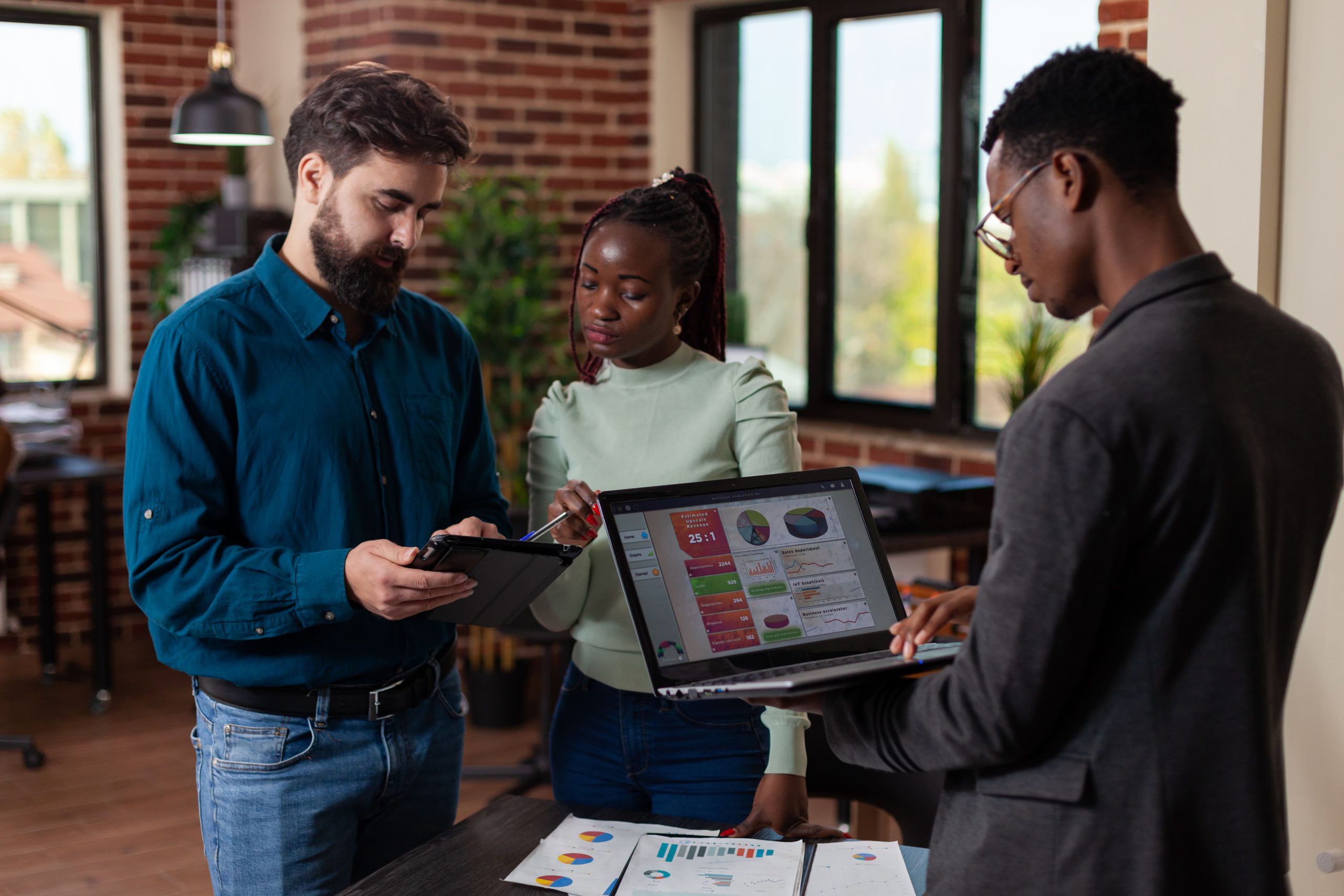 A diverse team of professionals discussing data on tablets and a laptop in a modern office setting with brick walls. The screens display various charts and graphs, indicating a focus on analytics and performance metrics.