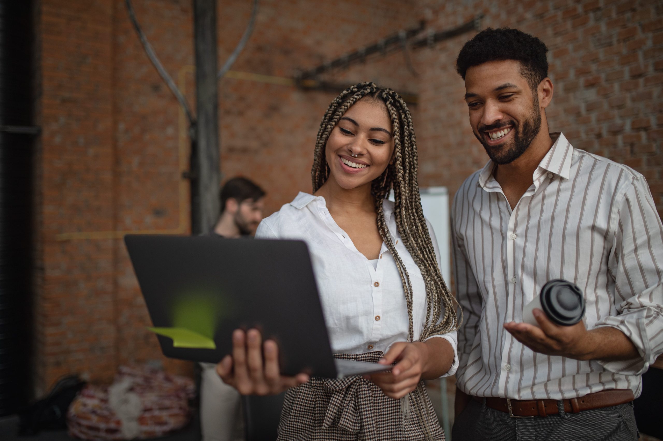 A cheerful businesswoman and a businessman reviewing something on a laptop screen while standing together in a modern workspace. The background shows exposed brick walls, adding to the urban office environment. The woman holds the laptop, and the man has a coffee cup, indicating a casual, collaborative discussion.