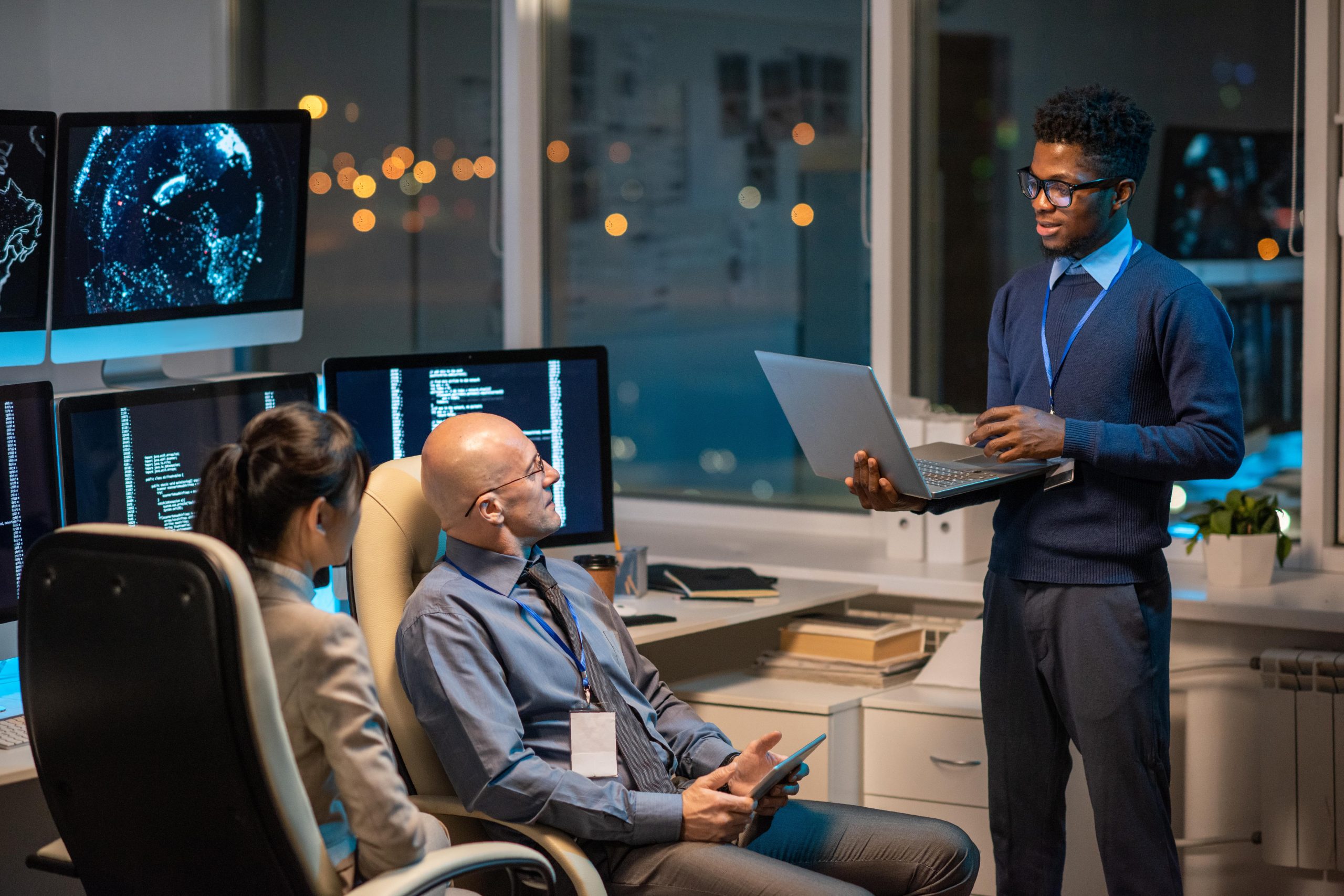 Young professional presents data on a laptop to colleagues in a modern office setting with screens displaying analytics.