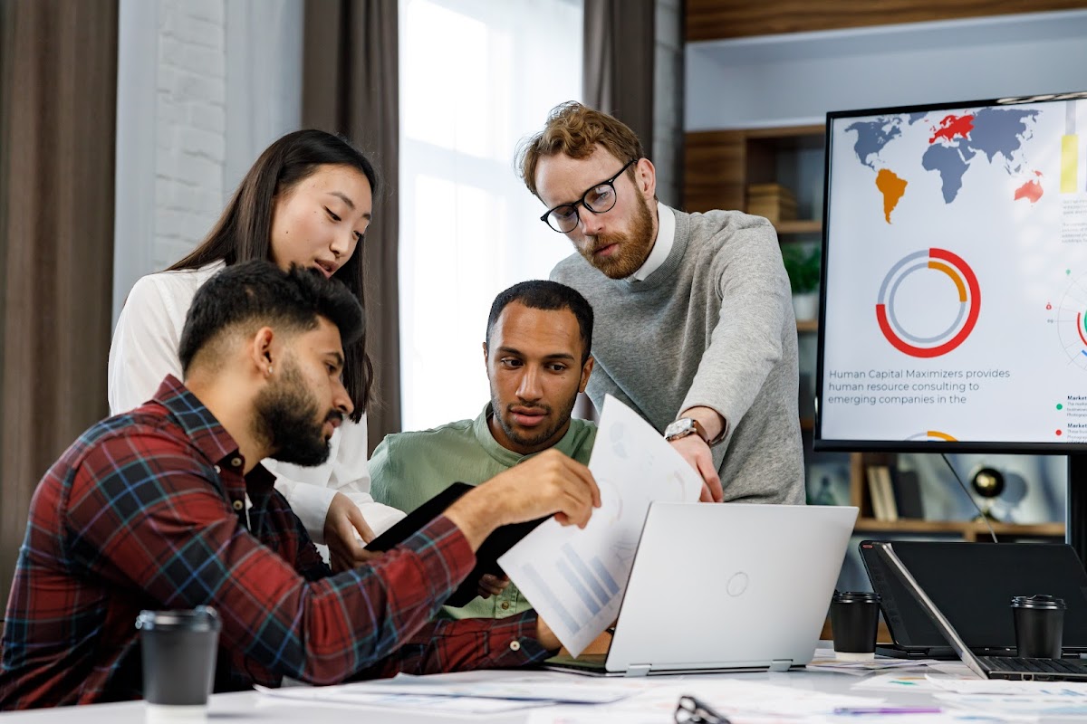 Diverse team collaborating over business documents with global data on screen.